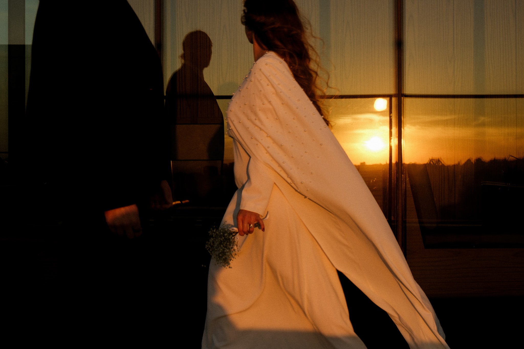 La mariée passe devant les baies vitrées au coucher du soleil, sa cape flottant derrière elle